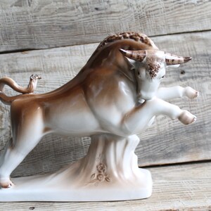 May include: A white and brown porcelain figurine of a bull standing on a base with a floral design. The bull is in a charging pose with its head lowered and its horns pointed forward.