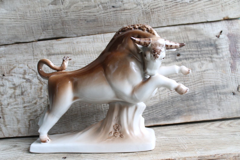 May include: A white and brown porcelain figurine of a bull standing on a base with a floral design. The bull is in a charging pose with its head lowered and its horns pointed forward.