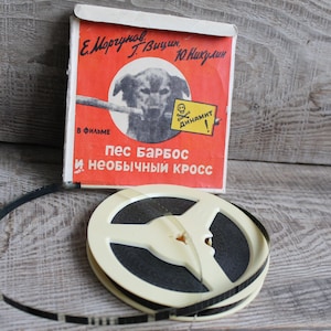 Puede incluir: Un carrete de película vintage con una tira de película en blanco y negro y un carrete de plástico blanco. El carrete está etiquetado con "Пес БАРБОС И НЕОБЫЧНЫЙ КРОСС" en escritura cirílica. La etiqueta también incluye los nombres "Е. Мориц", "Вицин", "Ю. Никулин", y "ДИНАМИТ!" en escritura cirílica.