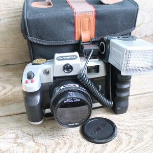 May include: A silver and black 35mm film camera with a black lens and a flash attached. The camera is sitting on a wooden surface with a black lens cap next to it.