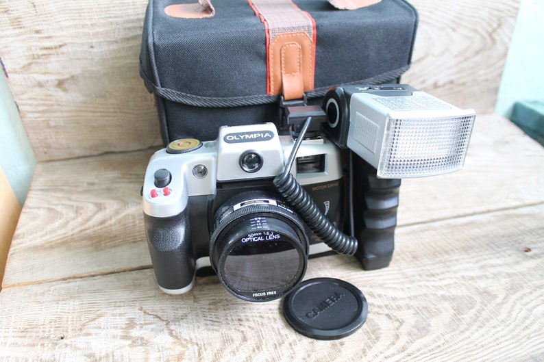 May include: A silver and black 35mm film camera with a black lens and a flash attached. The camera is sitting on a wooden surface with a black lens cap next to it.