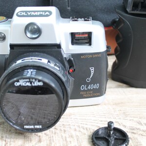 May include: A silver and black 35mm film camera with a black lens and the text "OLYMPIA OL4040 MOTOR DRIVE RED EYE REDUCTION" on the side.