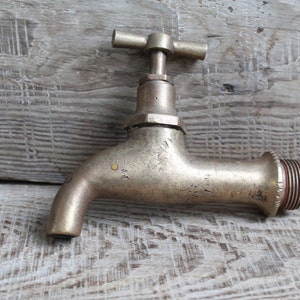 May include: A vintage-style brass faucet featuring a T-shaped handle at the top, a curved spout, and a threaded connection point on the side. The metal surface displays a weathered, matte finish with subtle signs of wear and a small yellow mark.