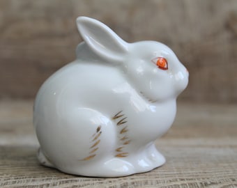 May include: A white porcelain rabbit figurine with orange eyes and gold accents. The rabbit is sitting with its head tilted down.
