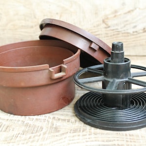 Może przedstawiać: Brązowy plastikowy zbiornik do wywoływania filmów z pokrywką i czarną szpulą filmową. Zbiornik jest cylindryczny z szerokim otworem. Szpula filmowa ma centralne wrzeciono i spiralny wzór.