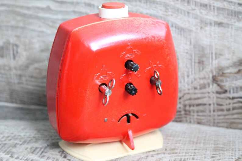 May include: A red wind-up alarm clock with three keys on the front. The clock has a white base and a red button on top.