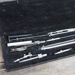 Può includere: Un set di compassi da disegno in una custodia di velluto nero. I compassi sono in metallo e hanno una finitura argentata. La custodia è foderata in velluto nero e ha una chiusura a scatto.