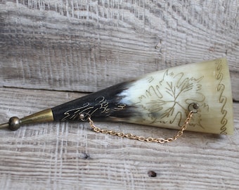 May include: A vintage horn-shaped container with a brass tip and chain. The container is cream-colored with a black base and features etched leaf designs and gold accents. The chain is gold-colored.