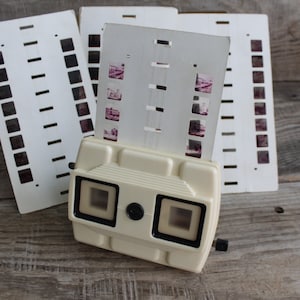 Puede incluir: Un visor 3D vintage de plástico blanco con dos ventanas de visualización rectangulares. El visor se utiliza para ver un conjunto de diapositivas de 35 mm, que están dispuestas en una tarjeta blanca con líneas negras.
