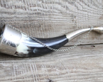 May include: A black and white horn drinking cup with a silver rim and chain. The horn is on a wooden surface.