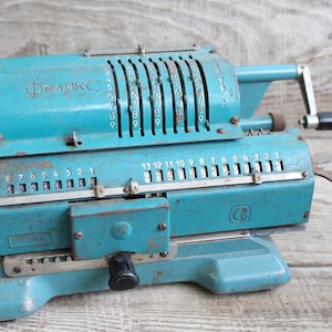 Puede incluir: Calculadora mecánica vintage de color turquesa con números visibles y manivela. La calculadora tiene la palabra cirílica "Феликс" en la parte superior. La calculadora es de metal y muestra signos de desgaste.