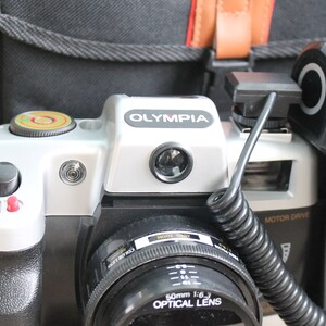 May include: A silver and black 35mm film camera with a black lens and a flash attached. The camera has the word "OLYMPIA" printed on the top.