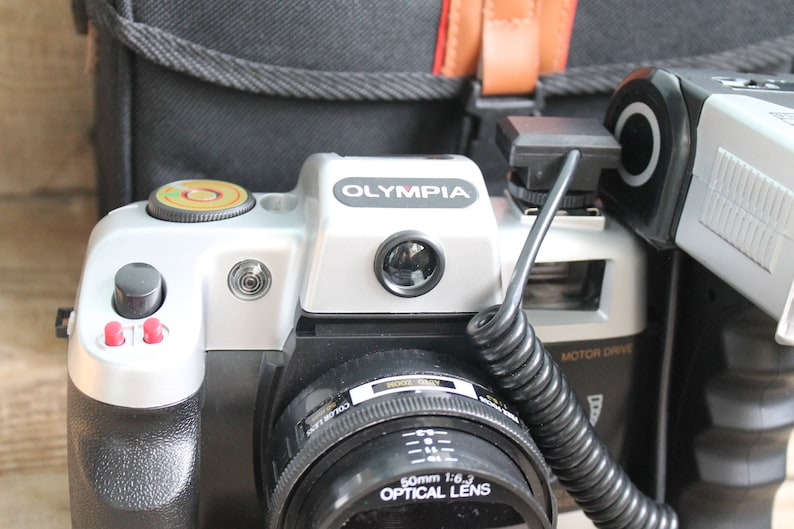 May include: A silver and black 35mm film camera with a black lens and a flash attached. The camera has the word "OLYMPIA" printed on the top.