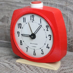 May include: A red alarm clock with a white face and black hands. The clock has a white button on top and a white base. The clock face has black numbers and the word "ЧacOBHK" in Cyrillic script.