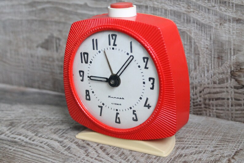 May include: A red alarm clock with a white face and black hands. The clock has a white button on top and a white base. The clock face has black numbers and the word "ЧacOBHK" in Cyrillic script.