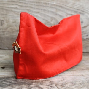 May include: Bright red cap with a star-shaped pin. The cap is made of a smooth, possibly satin-like material. The pin features a portrait and a red star. The cap is displayed on a wooden surface.