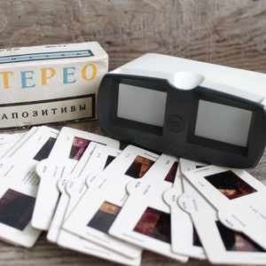 Puede incluir: Un visor estéreo vintage en blanco y negro, acompañado de una caja y un conjunto de diapositivas. La caja tiene la palabra "STEREO" en amarillo y el texto "ДИАПОЗИТИВЫ". Las diapositivas son blancas con marcos negros.