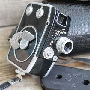 Puede incluir: Una cámara de cine vintage de 8 mm en negro y plata con el nombre "Kran" impreso en el lateral. La cámara está sobre una funda de cuero negro con estampado de cocodrilo.