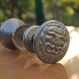 Könnte beinhalten: Ein Satz antiker, silberfarbener Türknöpfe. Der vordere Knopf zeigt ein detailliertes Relief eines bärtigen Gesichts. Die Knöpfe sind in einer Reihe angeordnet, wobei sich der Gesichts-Knopf im Vordergrund befindet. Der Hintergrund ist unscharf.