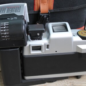 May include: A silver and black camera with a built-in flash. The camera has a black lens and a silver body with a black grip. The flash is attached to the top of the camera and has a black housing with a silver face.