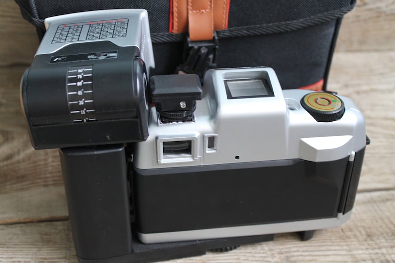 May include: A silver and black camera with a built-in flash. The camera has a black lens and a silver body with a black grip. The flash is attached to the top of the camera and has a black housing with a silver face.