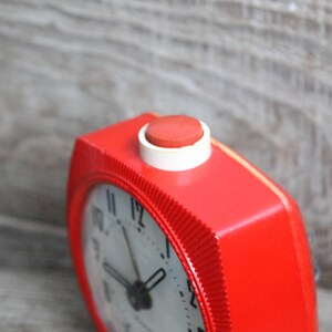 May include: A red alarm clock with a white face and black hands. The clock has a red button on the top.