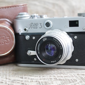 May include: A silver and black FED 3 rangefinder camera with a brown leather case. The camera body features the text FED 3 in Cyrillic and Latin characters. The lens is marked with Industar-25M, 1:2.8, and F=5cm, along with a serial number.
