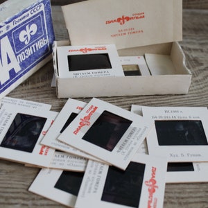 Puede incluir: Juego de diapositivas vintage con una caja azul y blanca y varias diapositivas esparcidas sobre una superficie de madera. La caja tiene texto cirílico y un logotipo. Las diapositivas están en marcos blancos con película negra.