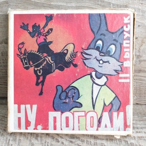 Puede incluir: Caja de cerillas vintage con una ilustración de dibujos animados de un conejo y un lobo a caballo sobre un fondo rojo. El texto "NU POGODI!" es visible. La caja tiene un aspecto desgastado.