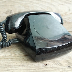 Puede incluir: Un teléfono negro vintage con un cable de auricular en espiral. El teléfono tiene un dial giratorio y un acabado brillante. El teléfono está sobre una superficie de madera clara.