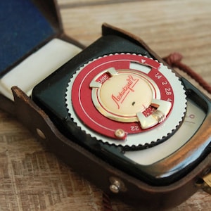 May include: A vintage light meter in a brown leather case. The meter has a red and silver dial with numbers and a gold-coloured centre. The case is open, revealing a blue interior.