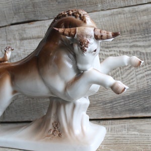May include: A white porcelain bull figurine with brown accents. The bull is standing on its hind legs with its front legs raised. The figurine is on a white base with brown floral details.