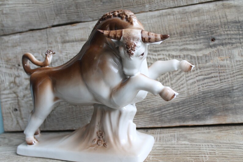May include: A white porcelain bull figurine with brown accents. The bull is standing on its hind legs with its front legs raised. The figurine is on a white base with brown floral details.