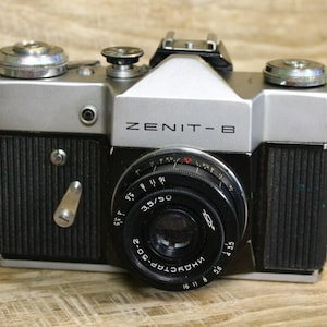 Puede incluir: Una cámara de película de 35 mm Zenit-B plateada y negra con un objetivo Industar-50-2 negro. La cámara tiene un cuerpo plateado con una cubierta de imitación de cuero negro y un objetivo negro con un anillo plateado.