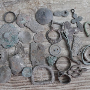 May include: A collection of antique metal artifacts, including buttons, rings, and decorative pieces, in various shapes and sizes. The items display a weathered, aged appearance with shades of gray, green, and brown, suggesting historical significance. The objects are arranged on a light-colored wooden surface.