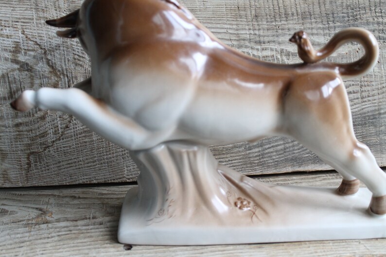 May include: A white and brown ceramic bull figurine with a small bird perched on its tail. The bull is standing on a base with a textured surface.
