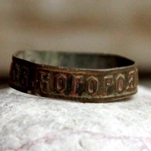 May include: A close-up of a weathered, bronze-colored ring with embossed lettering. The ring is set against a light-colored, textured background. The ring's surface shows signs of age and wear.