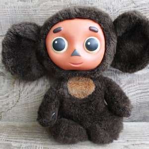 May include: A plush toy featuring a brown furry body and large, rounded ears. The toy has a light brown face with large, round eyes and a small nose. A patch of lighter brown fur is on the chest.