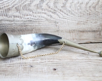 May include: A black and white horn drinking cup with a metal rim and a gold chain attached. The horn is on a wooden surface.