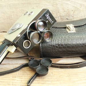 Puede incluir: Cámara de cine vintage Ekran-4 con cuerpo negro y gris, múltiples lentes y el texto "Ekran-4". Se acompaña de una funda de cuero negra con correa y un soporte para la tapa del objetivo. La cámara tiene un asa.