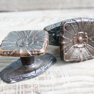Può includere: Pomelli per porte antichi in bronzo con un design floreale quadrato. I pomelli hanno una superficie dettagliata e strutturata e una patina scura e invecchiata. L'hardware è probabilmente vintage e adatto a progetti di restauro.