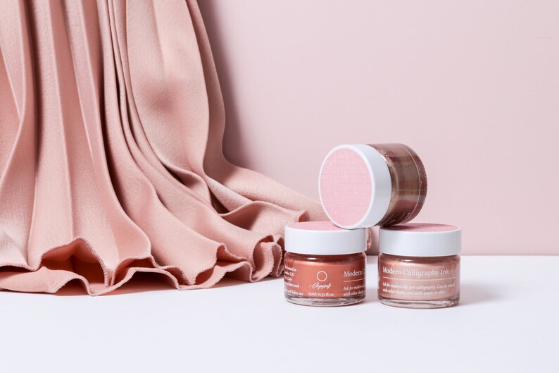 May include: Three jars of modern calligraphy ink in shades of pink and copper, with white lids and pink labels. The jars are stacked on a white surface, with a pink pleated fabric and a pink backdrop.