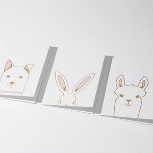 May include: Three white greeting cards with gold and pink animal illustrations: a dog, a rabbit, and a llama. The dog has a pink collar, the rabbit has long ears, and the llama has a smiling face. The cards are arranged in a row.