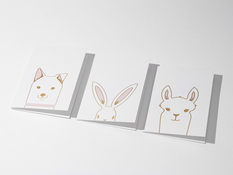 May include: Three white greeting cards with gold and pink animal illustrations: a dog, a rabbit, and a llama. The dog has a pink collar, the rabbit has long ears, and the llama has a smiling face. The cards are arranged in a row.