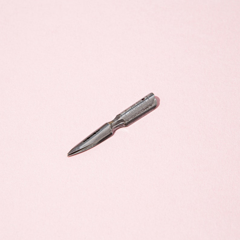 Pu&ograve; includere: Un pennino da calligrafia in metallo vintage con la scritta "ISERLOHN" incisa sul lato. Il pennino &egrave; di colore argento e si trova su uno sfondo rosa chiaro.