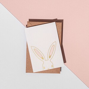 May include: A white greeting card with a gold bunny illustration and pink accents, resting on a brown envelope. The card and envelope are set against a backdrop of white and pink geometric shapes, creating a minimalist aesthetic.