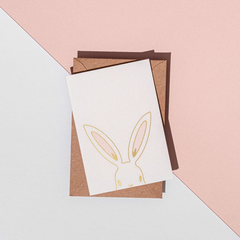 May include: A white greeting card with a gold bunny illustration and pink accents, resting on a brown envelope. The card and envelope are set against a backdrop of white and pink geometric shapes, creating a minimalist aesthetic.