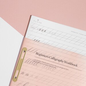 Pu&ograve; includere: Un quaderno di calligrafia con copertina rosa e foglio di pratica bianco. La copertina recita "Beginners Calligraphy Workbook". Il foglio di pratica ha linee a griglia e la parola "ALAISE" in alto. Una clip color oro tiene insieme le pagine.