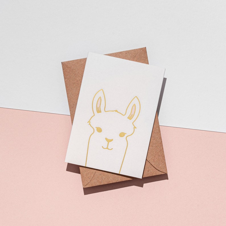 May include: A white greeting card with a gold outline illustration of a llama, resting on a brown envelope. The card and envelope are on a pink and white background. The llama illustration features a simple, minimalist design.