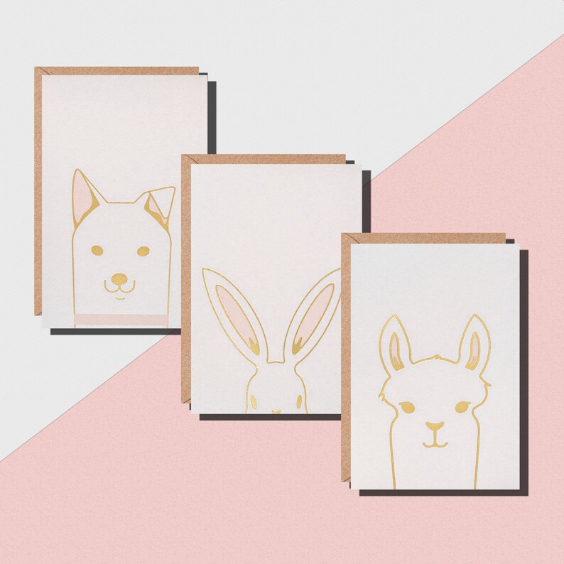 May include: Three greeting cards with gold foil animal illustrations: a dog, a rabbit, and a llama. Each card has a white background with a gold outline of the animal's face and ears. The cards have a brown border and are arranged on a pink and white surface.
